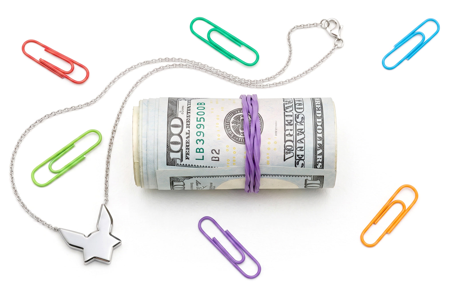 A roll of hundred-dollar notes held by a rubber band, lying among scattered coloured paperclips and a silver chain with a bird pendant